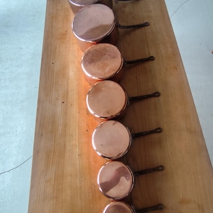 May include: A set of seven graduated copper saucepans arranged in a single line from largest to smallest. Each round saucepan features a polished metallic copper exterior and a dark, slender metal handle extending from the side of the vessel.