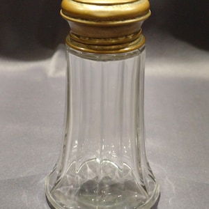 Op de afbeelding: Een heldere glazen pot met een messing deksel. De pot heeft een uitlopende basis en verticale geribbelde zijkanten. Het deksel heeft een afgeronde vorm met een decoratieve rand. De pot is leeg en lijkt antiek te zijn.