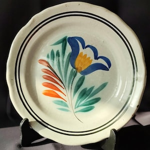Könnte beinhalten: Ein weißer Keramikteller mit handgemaltem Blumendesign. Der Teller hat eine blaue Blume mit gelbem Zentrum, grüne Blätter und orangefarbene Akzente. Er hat einen schwarzen, doppelt gefütterten Rand und einen gewellten Rand.