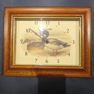 Może przedstawiać: Prostokątny zegar ścienny z brązową drewnianą ramą. Tarcza zegara przedstawia scenę przyrodniczą z dwoma kaczkami i napisem "BAYARD". Wskazówki zegara są czarne, a cyfry w klasycznej czcionce.