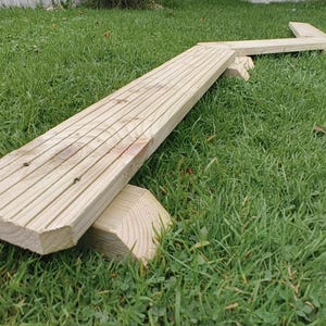 May include: A wooden balance beam, made from light-coloured wood, set on a grassy surface. The beam has a long, flat section with grooved lines and is supported by a wooden base. The structure is designed for outdoor play and measures approximately 150 cm in length.