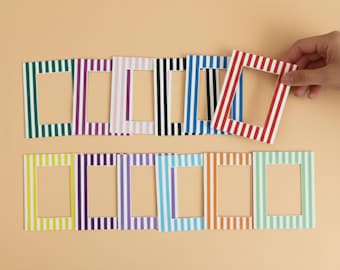 May include: A collection of colorful striped picture frames in various colors, including green, purple, white, black, blue, red, yellow, and orange. One frame is being held by a hand. The frames are rectangular with a central opening.