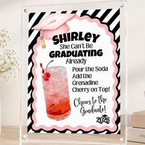 May include: A clear acrylic graduation sign with a pink and black border. The sign features a pink graduation cap and a cocktail illustration. Text includes "SHIRLEY She Can't Be GRADUATING Already" and "Cheers to the Graduate!" with the year 2026.