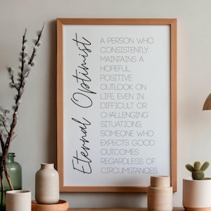 Op de afbeelding: Een ingelijste print met de woorden "Eternal Optimist" in een stijlvol lettertype. De tekst definieert een optimist. De print wordt getoond met decoratieve vazen, een kleine cactus en een hangende goudkleurige lamp.