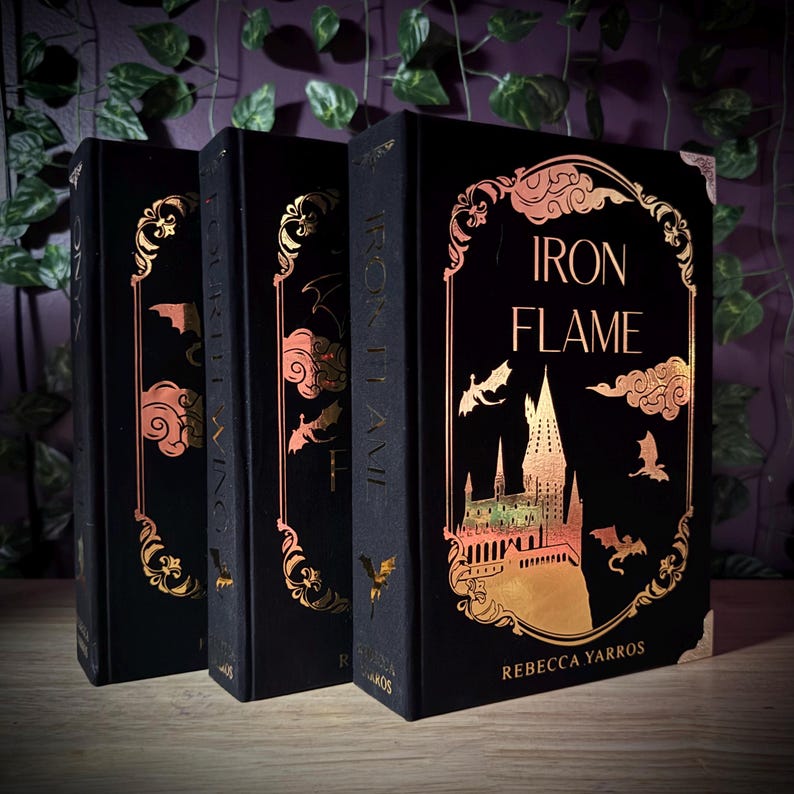 May include: Three hardcover books with black covers and gold foil accents. The book spines and covers feature intricate designs, including the title "Iron Flame" and the author's name. The books are displayed on a wooden surface.