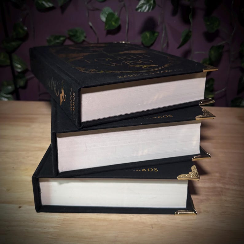 May include: A stack of three hardback books with black covers and white pages. The top book has gold lettering that reads "Fourth Wing" and "Rebecca Yarros". Each book has gold corner protectors. The books are stacked on a wooden surface.