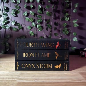 May include: Three hardcover books stacked on a wooden surface. The books are black with gold lettering and illustrations. Titles include "Fourth Wing," "Iron Flame," and "Onyx Storm." The author's name, Rebecca Yarros, is also visible.