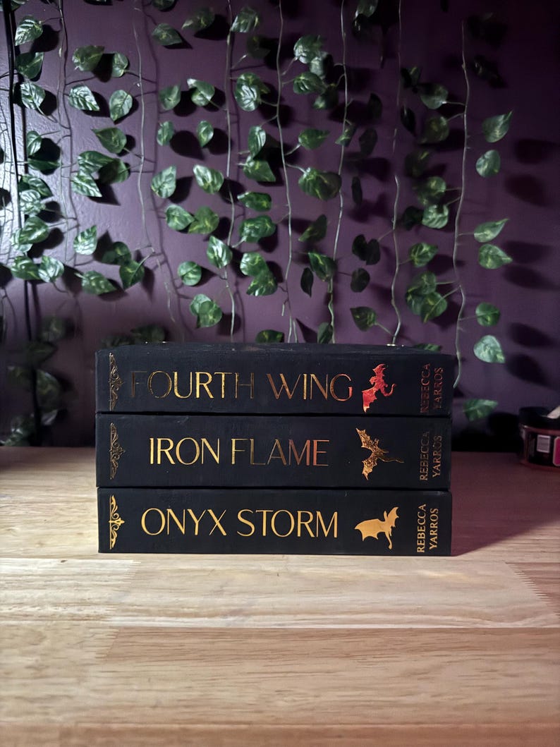 May include: Three hardcover books stacked on a wooden surface. The books are black with gold lettering and illustrations. Titles include "Fourth Wing," "Iron Flame," and "Onyx Storm." The author's name, Rebecca Yarros, is also visible.