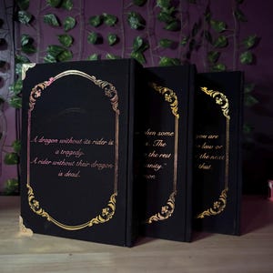 May include: Three black books with gold ornate frames and text. The first book reads, "A dragon without its rider is a tragedy. A rider without their dragon is dead." The other books have quotes. The books are on a wooden surface.