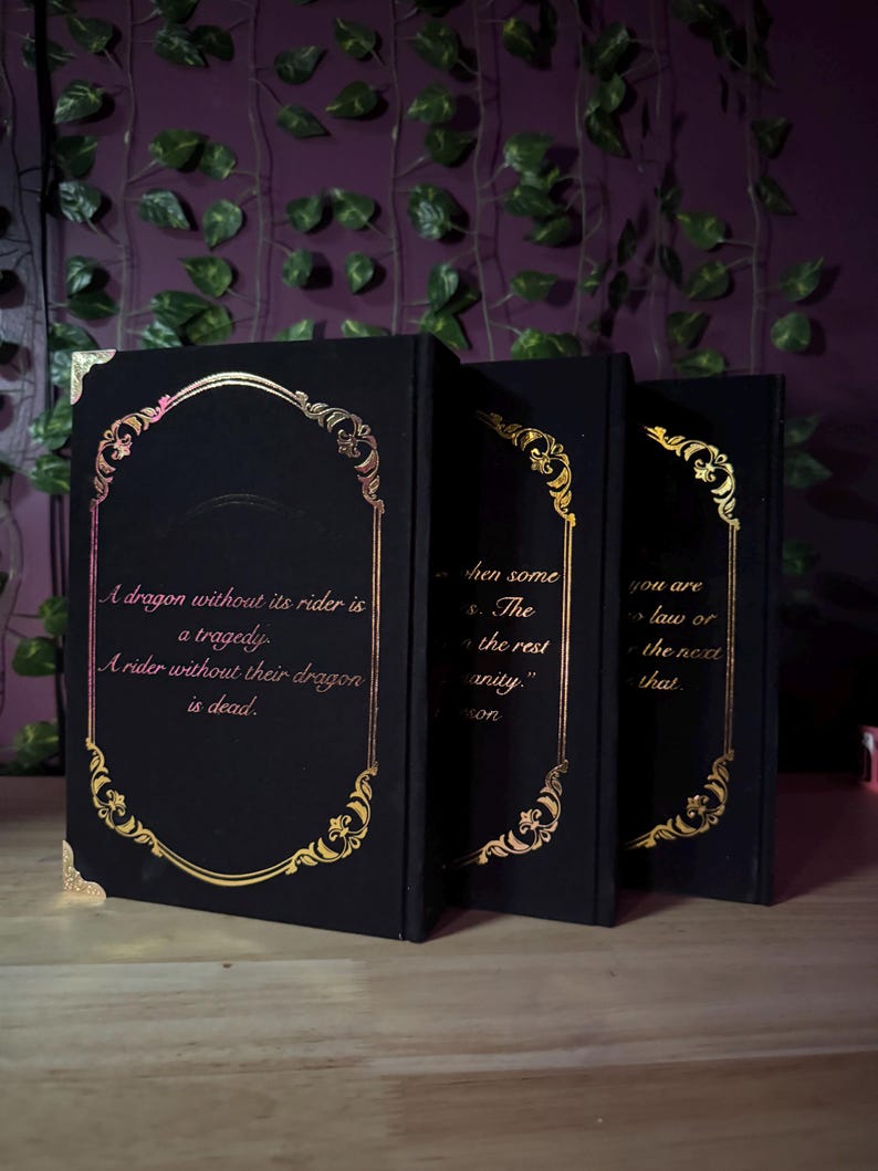 May include: Three black books with gold ornate frames and text. The first book reads, "A dragon without its rider is a tragedy. A rider without their dragon is dead." The other books have quotes. The books are on a wooden surface.