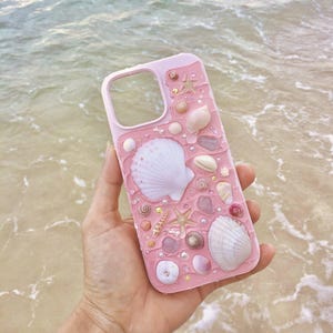 Puede incluir: Una funda de teléfono rosa decorada con conchas marinas, estrellas de mar y pequeñas cuentas. La funda tiene un acabado brillante y un recorte para la cámara. El fondo muestra el océano y la arena.