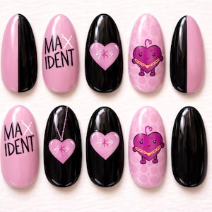 Puede incluir: Un conjunto de diez uñas postizas de forma ovalada. Los diseños incluyen negro liso, rosa con el texto "MAX IDENT", negro con un corazón rosa y rosa con un personaje de corazón de dibujos animados. Las uñas están dispuestas sobre una superficie blanca.
