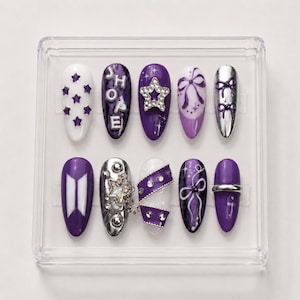May include: A set of ten press-on nails in a clear plastic case, featuring purple, white, and silver designs. Decorations include the text J-HOPE, a BTS logo, rhinestones, star shapes, bows, and metallic silver accents on almond-shaped nail tips.