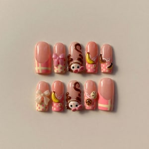 Könnte beinhalten: Ein Satz von zehn künstlichen Nägeln mit hellrosa Basis und verschiedenen Designs. Einige Nägel zeigen eine rosa Schleife, einen Affen oder eine Banane. Andere haben ein rosa-gelbes Karomuster oder Tupfenmuster.