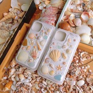 Może przedstawiać: Dwa przezroczyste etui na telefon ozdobione różnymi muszlami, rozgwiazdami i perłami. Etui mają różne kolory, w tym biały, beżowy i pastelowe odcienie. Etui otoczone są kolekcją muszli.