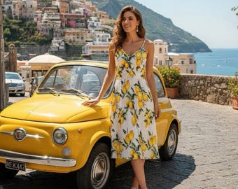 May include: A woman in a white dress with a yellow lemon print stands beside a bright yellow vintage car. The dress has spaghetti straps and a V-neck. The car is parked on a cobbled street with a scenic coastal background.