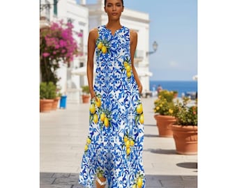 May include: A sleeveless, floor-length dress featuring a blue and white floral pattern with yellow lemons. The dress has a V-neck and pockets. The model is wearing white sandals. The background shows a sunny outdoor setting with buildings and potted plants.
