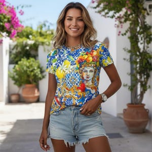 May include: A short-sleeved, crew neck t-shirt with a vibrant, ornate design featuring a woman's portrait, lemons, and fruit. The shirt is predominantly blue, yellow, and white. The model is wearing denim shorts and a watch.