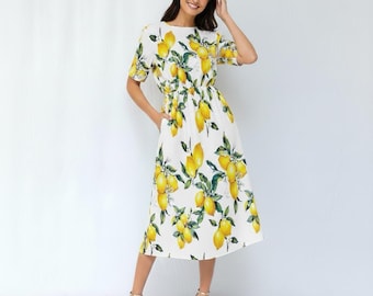 May include: A white midi dress featuring a vibrant lemon print. The dress has short sleeves, a fitted waist, and a flowing skirt. The yellow lemons are scattered across the fabric, with green leaves. The model is wearing gold heels.