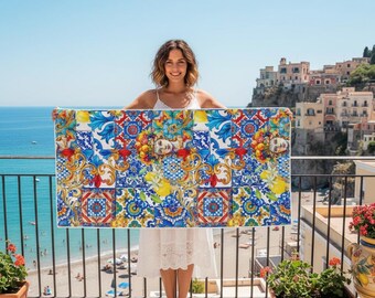 May include: A rectangular beach towel with a vibrant tile design. The towel displays a pattern of blue, yellow, red, and white squares with floral and face motifs. The towel is held up against a backdrop of the sea and a coastal town.