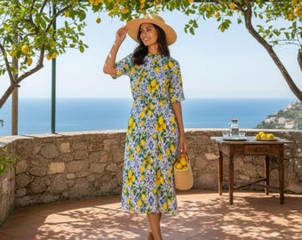 May include: A woman wearing a straw hat and a midi dress with a lemon print, standing under a lemon tree. The dress features a yellow and blue pattern. She is holding a woven basket with lemons. The background shows the sea and a small table.