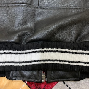 May include: Close-up of a black leather jacket with a ribbed knit hem. The hem features white stripes. A silver zipper is partially visible.
