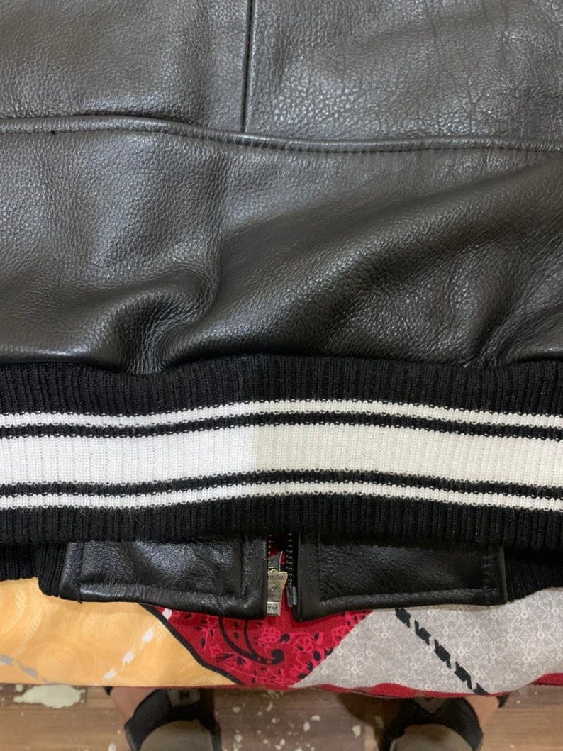 May include: Close-up of a black leather jacket with a ribbed knit hem. The hem features white stripes. A silver zipper is partially visible.