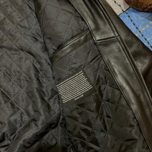 May include: Black leather jacket with a quilted black lining. The interior features a diamond-stitched pattern and a small rectangular tag with printed text. The jacket's exterior is smooth and shiny, with a zipper visible along the edge.