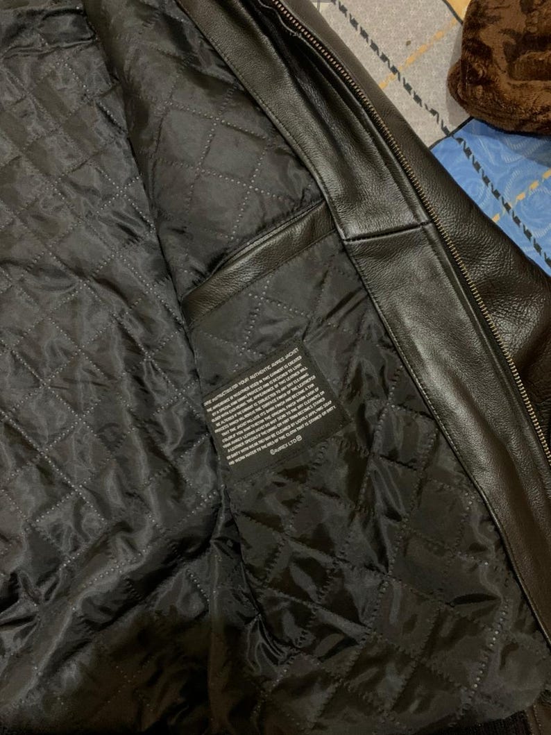 May include: Black leather jacket with a quilted black lining. The interior features a diamond-stitched pattern and a small rectangular tag with printed text. The jacket's exterior is smooth and shiny, with a zipper visible along the edge.