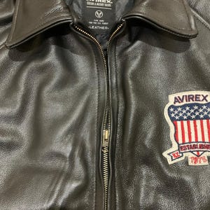 May include: Black leather jacket with a zipper closure and a collared neckline. The jacket features a patch on the right chest with the text "AVIREX" and "ESTABLISHED 1975". The jacket has a quilted lining and the text "AUTHENTIC 1975" is visible.