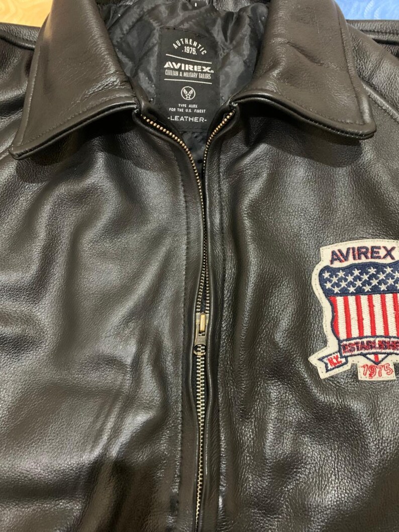 May include: Black leather jacket with a zipper closure and a collared neckline. The jacket features a patch on the right chest with the text "AVIREX" and "ESTABLISHED 1975". The jacket has a quilted lining and the text "AUTHENTIC 1975" is visible.