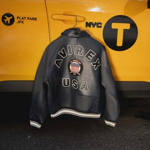 May include: A navy blue leather jacket with the word "AVIREX" and "USA" in white lettering on the back. The jacket has a patch with the words "All Americans" and a striped design. The jacket is hanging in front of a yellow taxi.