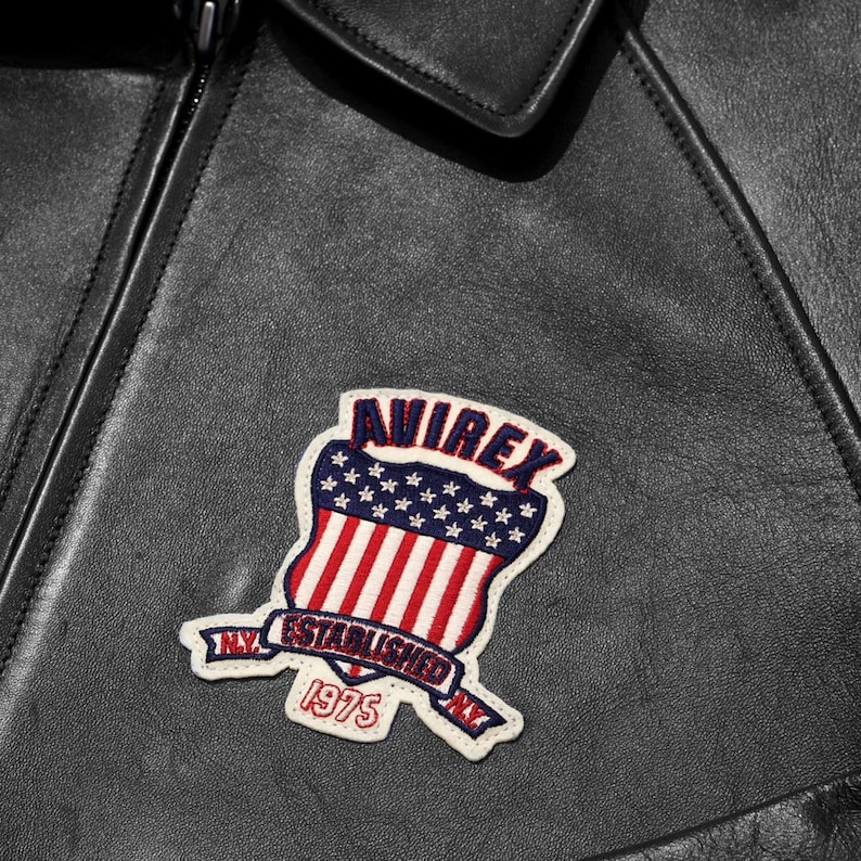 May include: Close-up of a black leather jacket with a patch. The patch is a shield design with red and white stripes, blue stars, and the text "AVIREX ESTABLISHED NY 1975". The jacket has a collar and a zipper.
