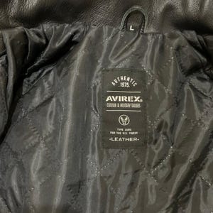 May include: Black leather jacket interior with a quilted lining. The jacket features a label with the text "AUTHENTIC 1975. AVIREX CIVILIAN & MILITARY TAILORS." The label also includes the text "TYPE AURK FOR THE U.S. FINEST -LEATHER-".