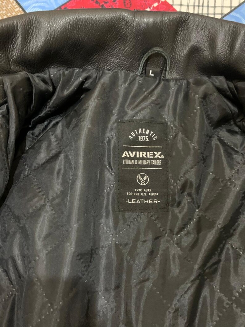 May include: Black leather jacket interior with a quilted lining. The jacket features a label with the text "AUTHENTIC 1975. AVIREX CIVILIAN & MILITARY TAILORS." The label also includes the text "TYPE AURK FOR THE U.S. FINEST -LEATHER-".