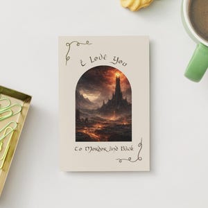 May include: A greeting card with the text "I Love You To Mordor and Back" features a fantasy landscape illustration within an arched frame. The artwork depicts a dark tower and fiery terrain. The card is on a white surface with a green-handled mug and a gold tray.