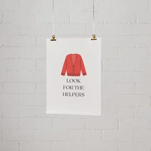 May include: A poster featuring a red cardigan illustration and the text "LOOK FOR THE HELPERS" in black. The poster is suspended against a white brick wall with gold clips.