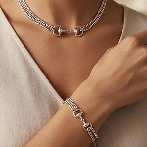 May include: A silver-toned jewelry set consisting of a double-chain choker necklace and a matching double-chain bracelet. Both pieces feature a central metal horsebit-style connector link that joins the two parallel curb chains together.