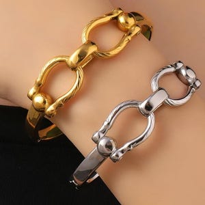 May include: Two hinged bangle bracelets, one in a polished gold tone and one in a polished silver tone. Each bracelet features a central horseshoe-shaped link with textured sides and a smooth, rounded ball accent at the top of the U-shape.