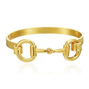 May include: A polished gold-tone bangle bracelet featuring a central horse bit motif. The design consists of a smooth, curved band connected to two circular rings joined by a horizontal bar, creating a continuous, reflective metallic surface.