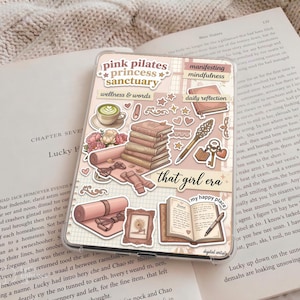 May include: A phone case with a pink and beige design, featuring stickers with text like "pink pilates princess sanctuary" and "that girl era." The case includes illustrations of books, a yoga mat, and a coffee cup.