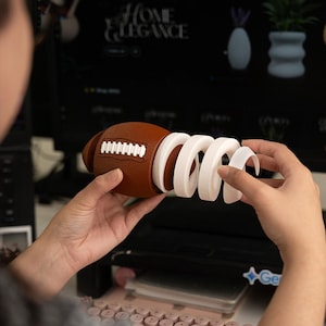 Peut inclure: Un ballon de football marron avec des coutures blanches est tenu avec un objet blanc en forme de spirale. Le ballon est fait d'un matériau texturé. L'arrière-plan comprend un écran d'ordinateur avec le texte "HOME ELEGANCE".