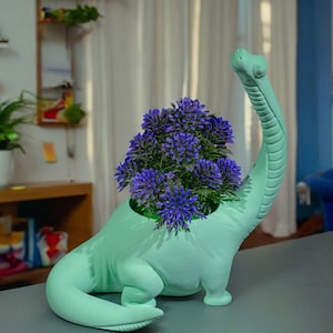 Puede incluir: Una maceta con forma de dinosaurio verde menta llena de vibrantes flores moradas. La maceta tiene un cuello largo y una superficie texturizada detallada. Las flores son de un tono morado intenso, creando un contraste llamativo.