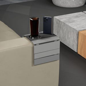 Puede incluir: Un reposabrazos de sofá gris multifuncional con un portavasos y una ranura para teléfono integrados. Un vaso de líquido oscuro y un teléfono inteligente negro están en el soporte. El reposabrazos está sujeto a un sofá beige.