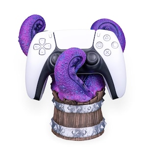 Könnte beinhalten: Ein weiß-schwarzer PlayStation 5-Controller wird von lila Tentakeln gehalten, die aus einem Holzfass kommen. Das Fass hat silberne Metallbänder und Nieten. Die Tentakel haben ein strukturiertes Aussehen.