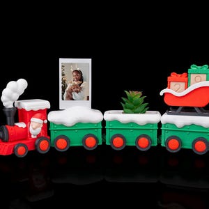 May include: A festive Christmas train set with a red engine and green cars. The train features a Santa Claus figure, a photo frame, a small succulent, and a sleigh carrying gifts. The train cars have a snow-like trim and red wheels.