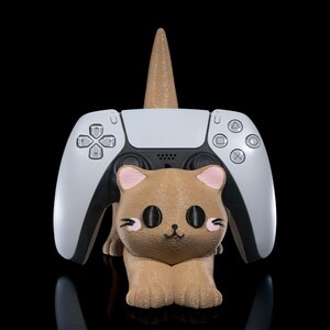 May include: A brown cat-shaped controller holder with pink accents, supporting a white and black game controller. A brown cone shape rises from the cat's head. The background is black.