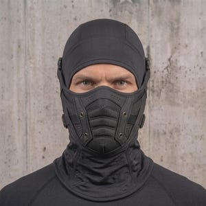 Op de afbeelding: Een zwarte tactische bivakmuts met een gestructureerd, meerdelig gezichtsmasker met geïntegreerde ventilatiegaten, versterkte zijbanden en metalen klinknagels. Het hoofddeksel bedekt het hoofd, de nek en de neus voor een volledige bescherming.