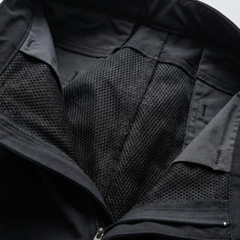 May include: Black trousers featuring a solid fabric exterior and a black mesh lining visible at the open waist area. The garment includes a zippered fly closure, reinforced stitching along the seams, and a structured waistband with belt loops.