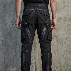 May include: Black tactical trousers featuring multiple utility pockets, integrated adjustable straps, and harness-style webbing. The pants include waterproof-style zippers on the thighs and lower legs, with additional side cargo panels and buckle closures.